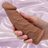 A hand with pink-painted nails holds the CalExotics Performance Maxx Rechargeable Vibrating Extension-Brown 7" against a lavender satin background.